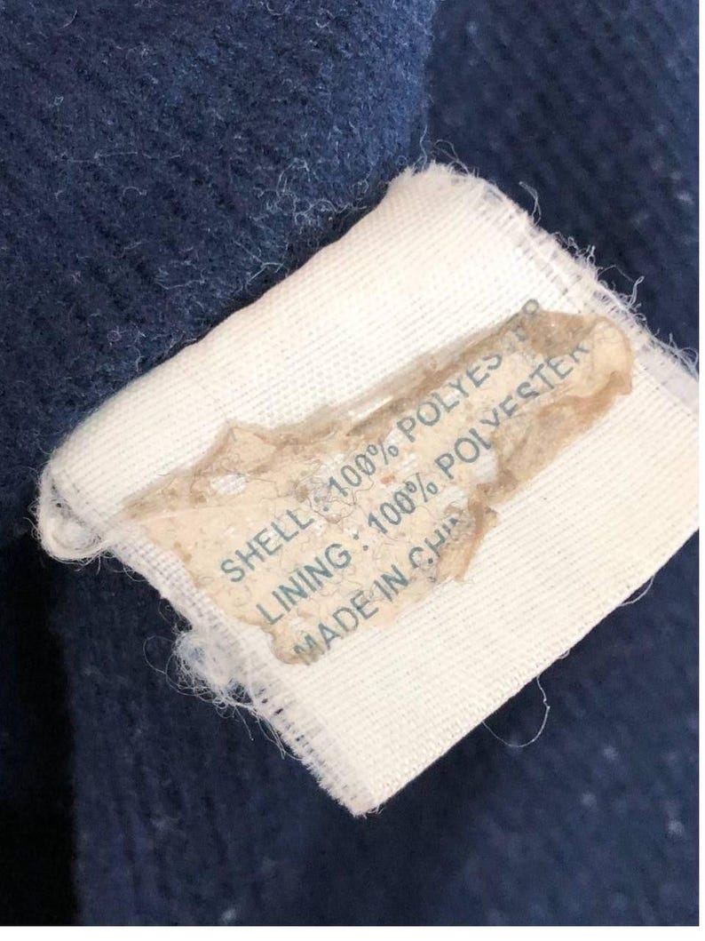 Puede incluir: Primer plano de una etiqueta sobre una tela azul marino. La etiqueta es blanca con una secci&oacute;n beige. El texto de la etiqueta dice "SHELL: 100% POLYESTER", "LINING: 100% POLYESTER" y "MADE IN CHINA".