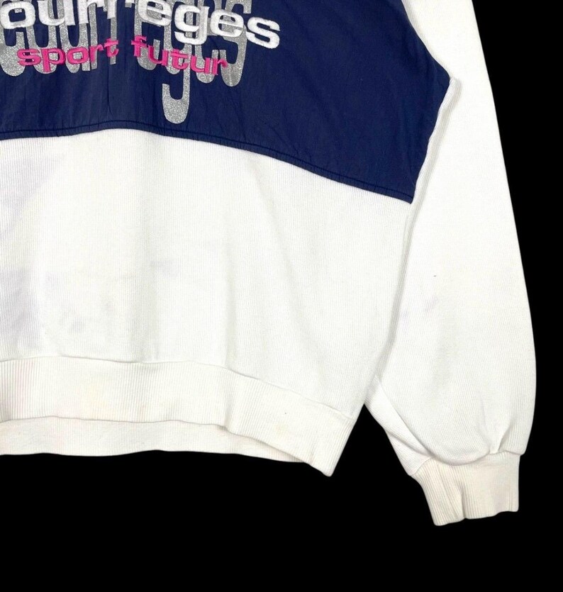 Puede incluir: Sudadera blanca con un panel azul marino en el pecho. El panel presenta el texto "courreges sport futur" en letras plateadas y rosas. La sudadera tiene pu&ntilde;os y dobladillo de canal&eacute;.