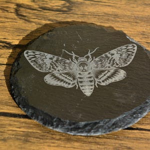 Puede incluir: Posavasos redondo de pizarra gris oscuro con un diseño grabado detallado de una polilla. La polilla tiene intrincados patrones de alas y está centrada en el posavasos. El posavasos está sobre una superficie de madera.
