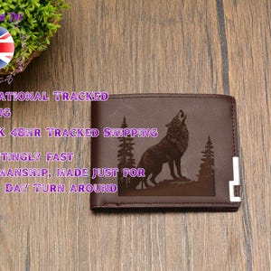May include: A brown leather wallet with a laser engraved image of a wolf howling at the moon. The wolf is surrounded by trees and the wallet has a silver metal trim.
