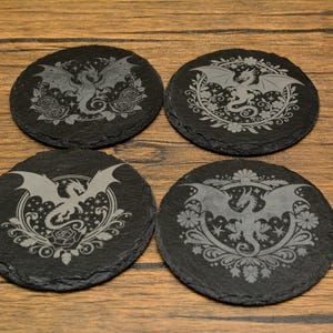 May include: Four black slate coasters with dragon designs. Each coaster features a different dragon illustration, surrounded by floral and decorative elements. The coasters are round and sit on a wooden surface.