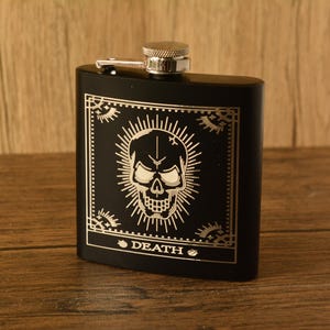 May include: Black hip flask with a silver cap and hinge. The front has an engraved skull design within a decorative frame, with the word "DEATH" below. The flask is on a wooden surface.