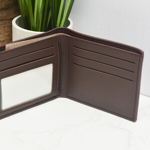 May include: A brown leather wallet with multiple card slots and a clear ID window. The wallet is open and laid flat on a white surface.