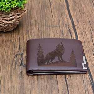 May include: A brown leather wallet with a wolf howling at the moon design engraved on the front. The wolf is surrounded by trees.