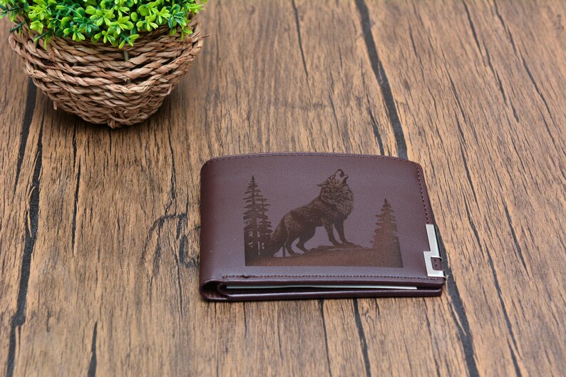 May include: A brown leather wallet with a wolf howling at the moon design engraved on the front. The wolf is surrounded by trees.