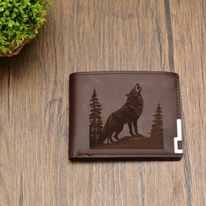 May include: Brown leather wallet with a wolf howling at the moon design. The wolf is surrounded by trees and the wallet has a silver metal accent.