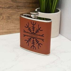 Pode incluir: Uma garrafa de couro marrom com tampa prateada e um design preto de um símbolo viking com a palavra "Dachshund" inscrita em uma faixa.