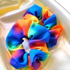 May include: Two rainbow-colored scrunchies made of soft fabric. The scrunchies are in a pile on a cream-colored satin surface.
