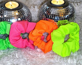 Könnte beinhalten: Vier bunte Haargummis in Grün, Pink, Orange und Limettengrün. Die Haargummis sind in einer Reihe auf einer weißen, flauschigen Oberfläche angeordnet. Im Hintergrund befinden sich zwei silberne Discokugel-Kerzenhalter.
