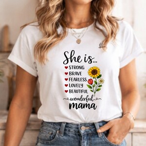 May include: White t-shirt with the text "She is..." and a floral design. The design includes a sunflower, red tulip, and small white flowers. The words "Strong, Brave, Fearless, Lovely, Beautiful, wonderful mama" are also printed on the shirt.
