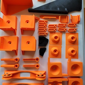 May include: An assortment of orange 3D-printed parts, including brackets, rollers, and clips, along with a black angled piece. The components are arranged on a white surface, showcasing various shapes and sizes, ideal for a DIY project.