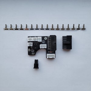 Op de afbeelding: Een zwarte printplaat met witte connectoren en een zwarte connector aan de zijkant. Er is een zwarte connector aan de rechterkant van de afbeelding. Er is een zilveren metalen strip met 18 kleine connectoren aan de bovenkant van de afbeelding. Er is een kleine zwarte connector aan de onderkant van de afbeelding.