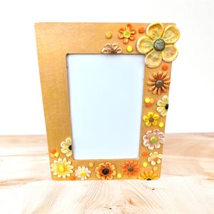 May include: Wooden photo frame with a light brown finish, adorned with colorful, three-dimensional floral embellishments in shades of yellow, orange, and brown. The frame has a rectangular opening for a photo.