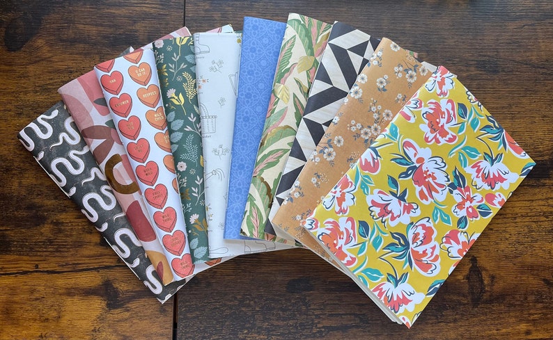 May include: A collection of nine rectangular sheets of patterned paper arranged in a fan shape. The designs include black and white wavy lines, pink and brown shapes, red hearts with text, green foliage, white line art, blue geometric patterns, tropical leaves, black and white triangles, tan floral print, and a yellow floral pattern.