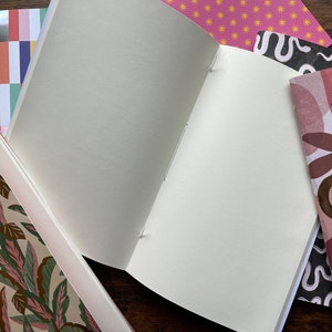 May include: A collection of notebooks with various patterned covers, including a green and pink leaf print, a multi-colored block stripe, a pink sun pattern, a black and white wavy line design, and a pink and brown abstract shape print with one open notebook.