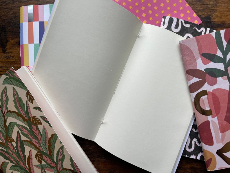 May include: A collection of notebooks with various patterned covers, including a green and pink leaf print, a multi-colored block stripe, a pink sun pattern, a black and white wavy line design, and a pink and brown abstract shape print with one open notebook.