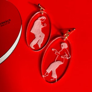 May include: A pair of clear oval-shaped earrings with a white silhouette of a person dancing inside each earring.