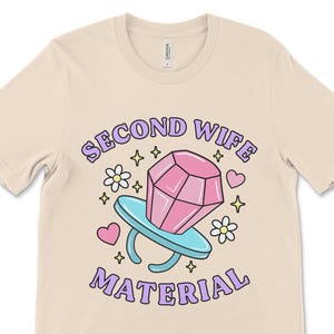 K&ouml;nnte beinhalten: Ein beiges T-Shirt mit dem Text "Second Wife Material" in lila Schrift. Ein Ring aus einer Zeichentrickfigur mit einem rosafarbenen Diamanten befindet sich in der Mitte des Textes. Der Ring ist von Herzen und Sternen umgeben.