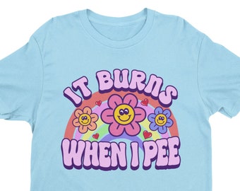 May include: A light blue t-shirt with a rainbow and flowers graphic. The text "IT BURNS WHEN I PEE" is printed in purple on the shirt.