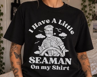 May include: A black short-sleeve t-shirt featuring a white graphic of a bearded sailor wearing a captain's hat and smoking a pipe while steering a ship. The text reads I Have A Little SEAMAN On my Shirt in a bold, arched white font.