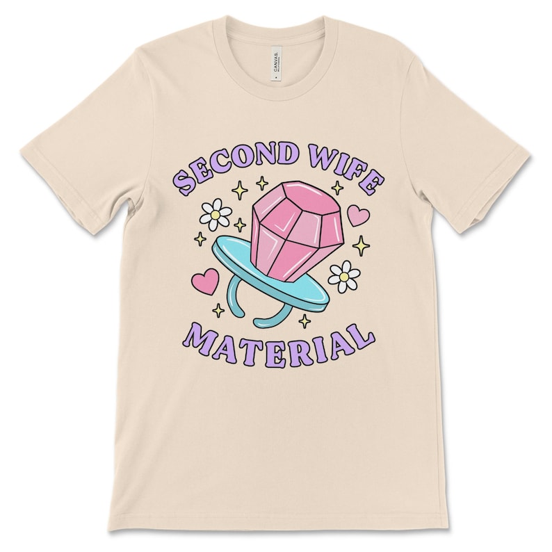 K&ouml;nnte beinhalten: Ein beiges T-Shirt mit dem Text "Second Wife Material" in lila Schrift. Ein Ring aus einer Zeichentrickfigur mit einem rosafarbenen Diamanten befindet sich in der Mitte des Textes. Der Ring ist von Herzen und Sternen umgeben.
