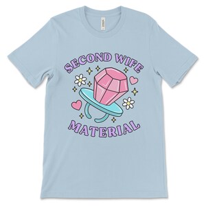 K&ouml;nnte beinhalten: Hellblaues T-Shirt mit einer Cartoon-Illustration eines Diamantrings mit dem Text "Second Wife Material" in Lila.