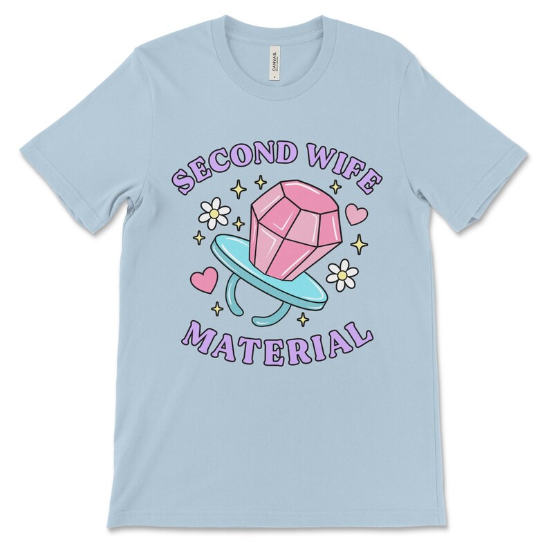 K&ouml;nnte beinhalten: Hellblaues T-Shirt mit einer Cartoon-Illustration eines Diamantrings mit dem Text "Second Wife Material" in Lila.