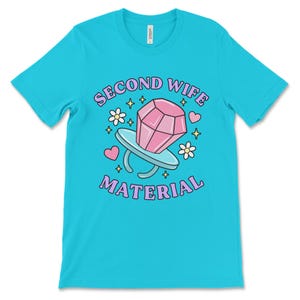 K&ouml;nnte beinhalten: Ein t&uuml;rkisfarbenes T-Shirt mit dem Text "Second Wife Material" in lila Schrift. Ein rosafarbener Diamantring mit einem rosafarbenen Stein befindet sich in der Mitte des Textes. Das Design ist von kleinen rosa Herzen, Sternen und Blumen umgeben.