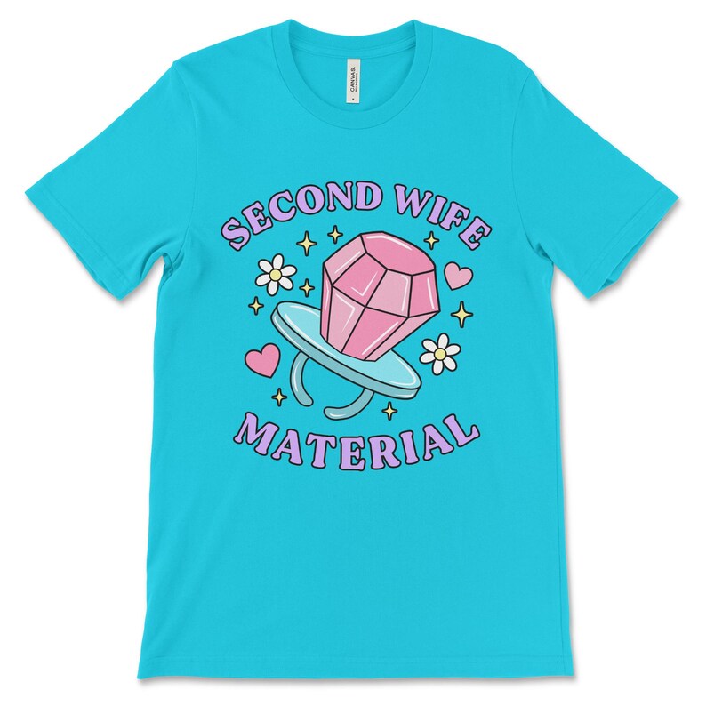 K&ouml;nnte beinhalten: Ein t&uuml;rkisfarbenes T-Shirt mit dem Text "Second Wife Material" in lila Schrift. Ein rosafarbener Diamantring mit einem rosafarbenen Stein befindet sich in der Mitte des Textes. Das Design ist von kleinen rosa Herzen, Sternen und Blumen umgeben.