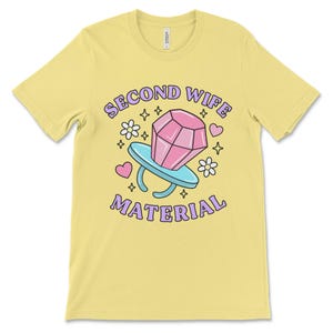 K&ouml;nnte beinhalten: Gelbes T-Shirt mit einer lila und pinkfarbenen Grafik eines Diamantrings mit dem Text "Second Wife Material" in lila.