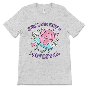 K&ouml;nnte beinhalten: Ein heidefarbenes T-Shirt mit einer lila und pinkfarbenen Grafik eines Diamantrings mit dem Text "Second Wife Material".