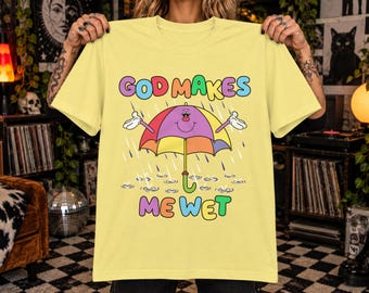 May include: A pale yellow t-shirt featuring a colorful graphic of a smiling umbrella character in the rain. The text "GOD MAKES ME WET" is printed in multi-colored bubble letters above and below the umbrella, which has white gloved hands.