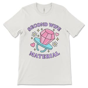 K&ouml;nnte beinhalten: Wei&szlig;es T-Shirt mit einem lila und pinkfarbenen Grafikdesign, auf dem "Second Wife Material" mit einem Cartoon-Diamantring und Herzen steht.