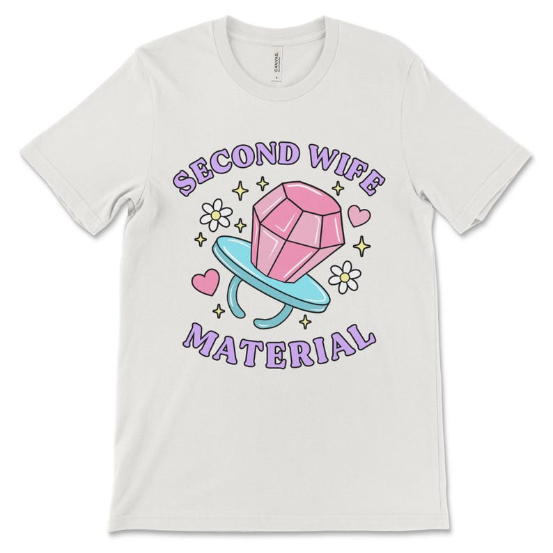 K&ouml;nnte beinhalten: Wei&szlig;es T-Shirt mit einem lila und pinkfarbenen Grafikdesign, auf dem "Second Wife Material" mit einem Cartoon-Diamantring und Herzen steht.