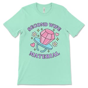 K&ouml;nnte beinhalten: Ein hellgr&uuml;nes T-Shirt mit dem Text "Second Wife Material" in lila Schrift. Ein Cartoon-Ring mit einem rosafarbenen Diamanten befindet sich in der Mitte des Textes. Der Ring ist von rosa Herzen, wei&szlig;en Blumen und goldenen Sternen umgeben.