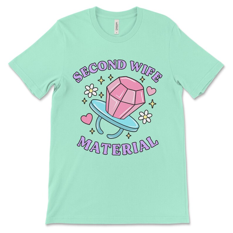 K&ouml;nnte beinhalten: Ein hellgr&uuml;nes T-Shirt mit dem Text "Second Wife Material" in lila Schrift. Ein Cartoon-Ring mit einem rosafarbenen Diamanten befindet sich in der Mitte des Textes. Der Ring ist von rosa Herzen, wei&szlig;en Blumen und goldenen Sternen umgeben.