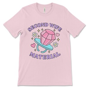K&ouml;nnte beinhalten: Pinkes T-Shirt mit einer Cartoon-Illustration eines Diamantrings mit dem Text "Second Wife Material" in lila Buchstaben.