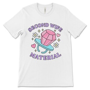 K&ouml;nnte beinhalten: Wei&szlig;es T-Shirt mit einem pinkfarbenen Diamant-Ring-Motiv und dem Text "Second Wife Material" in lila Schrift.