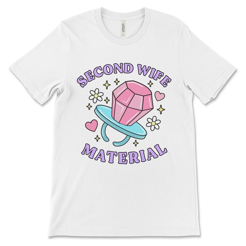 K&ouml;nnte beinhalten: Wei&szlig;es T-Shirt mit einem pinkfarbenen Diamant-Ring-Motiv und dem Text "Second Wife Material" in lila Schrift.