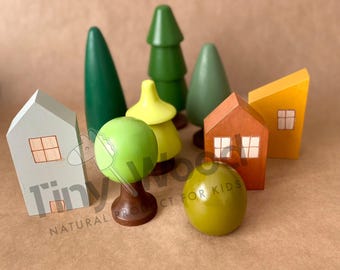May include: A collection of wooden toy houses, trees, and a round object. The houses are painted white, orange, and yellow, with small square windows. The trees are green, and the round object is light green. The text "Tiny Wood" and "Natural Product for Kids" are visible.
