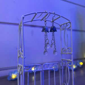 May include: Silver earrings with a bell-shaped charm and dangling crystal beads. The earrings are displayed on a white metal stand.