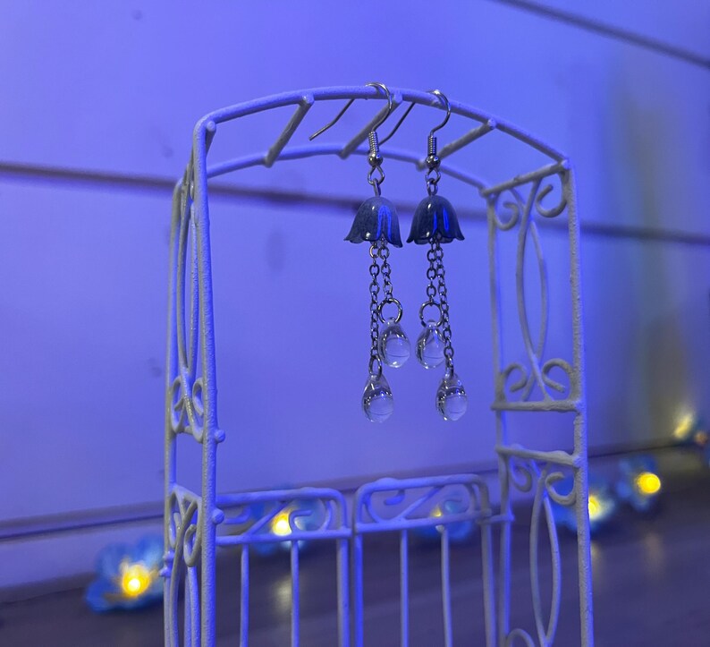 May include: Silver earrings with a bell-shaped charm and dangling crystal beads. The earrings are displayed on a white metal stand.
