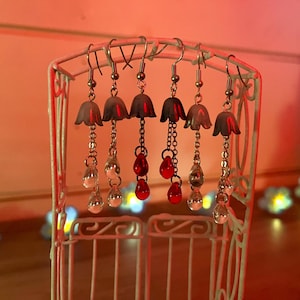 May include: Five pairs of silver dangle earrings with white bell-shaped charms and red glass beads hanging from them. The earrings are displayed on a white metal stand.