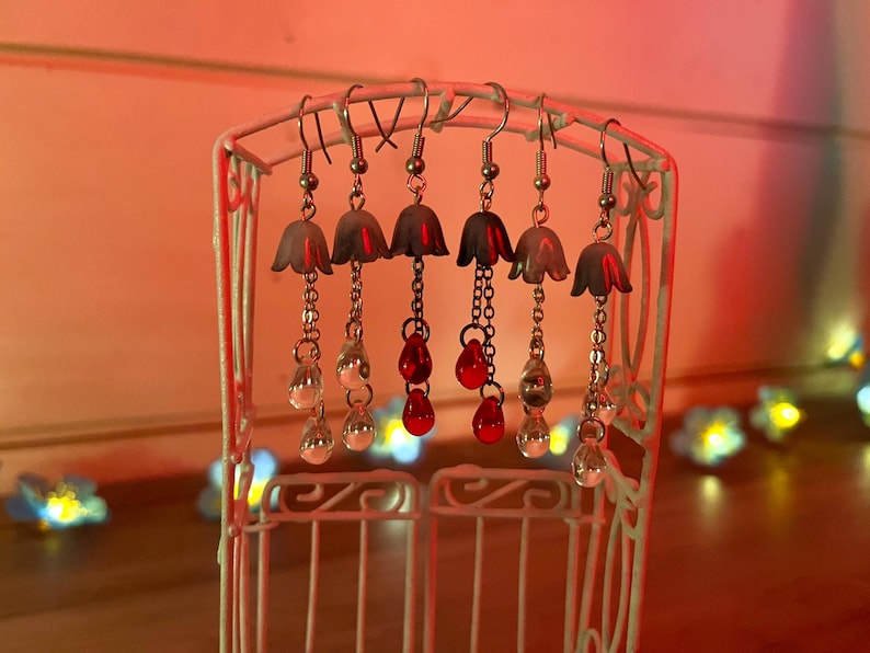 May include: Five pairs of silver dangle earrings with white bell-shaped charms and red glass beads hanging from them. The earrings are displayed on a white metal stand.