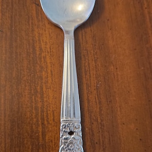 May include: A silver spoon with a rounded bowl and a decorative handle. The handle features vertical lines and an ornate floral design. The spoon is made of a shiny, reflective metal.