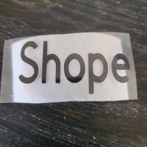 May include: A rectangular sticker with the word "Shope" in a bold, black font. The sticker is on a clear, transparent backing, and the background is a dark, textured surface.