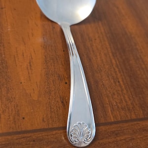 May include: A silver soup ladle with a rounded bowl and a long, curved handle. The handle has a decorative floral design at the end. The ladle is made of shiny metal and reflects the surrounding environment.