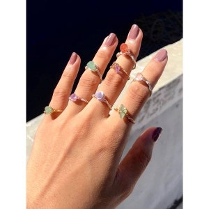 May include: A hand wearing several rings with different colored stones. The rings are made of silver wire and feature various shapes of stones, including round, oval, and irregular shapes. The stones are in shades of green, pink, purple, and orange.