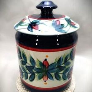 May include: A cylindrical ceramic canister with a matching lid featuring a dark blue knob. The body and lid are decorated with a hand-painted pattern of green leaves, blue petals, and red berries on a cream background, framed by red and blue bands.