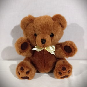 May include: A brown teddy bear with a yellow bow sitting on a white surface. The bear has black eyes and a black nose.